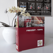 Individuell bedruckter Ordner in Rot mit der Aufschrift Freizeit-Tipps, dekorativ auf einem Tisch